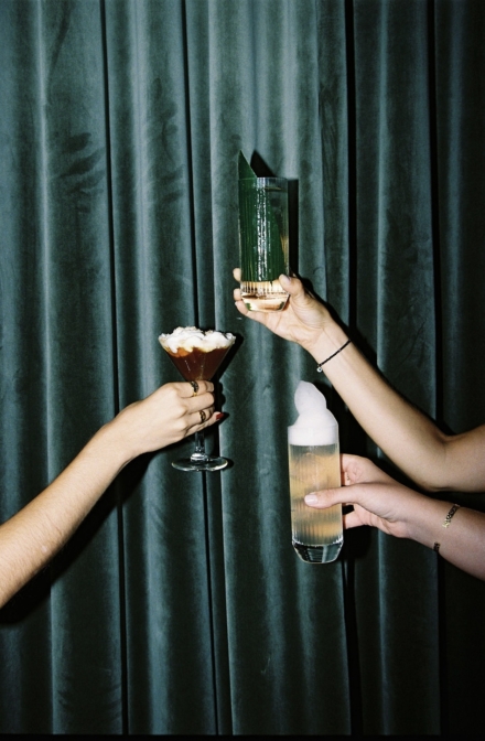 Dos manos brindan con un espresso martini y un cóctel verde frente a una cortina de terciopelo verde